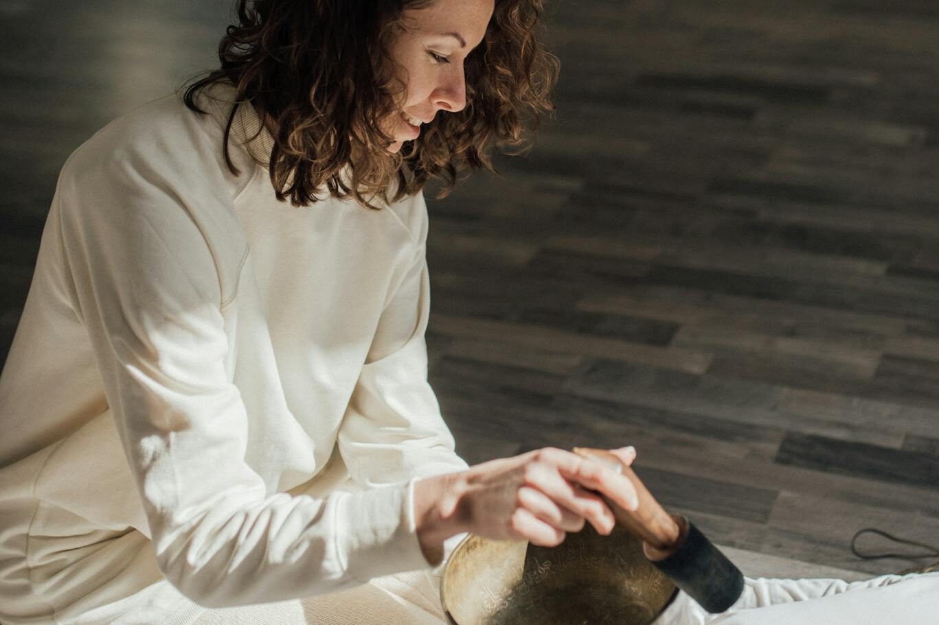 A woman performs sound therapy with a Tibetan singing bowl during a meditation session indoors.